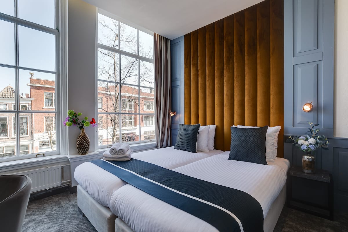 A hotel room with a large window, a bed with white and dark blue bedding, a wooden headboard, and a small table with a vase of flowers.