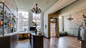 A hotel lobby with a reception desk, a seating area with a sofa and chairs, and a large window offering a view of the city. - Hotel Royal Bridges (Delft)