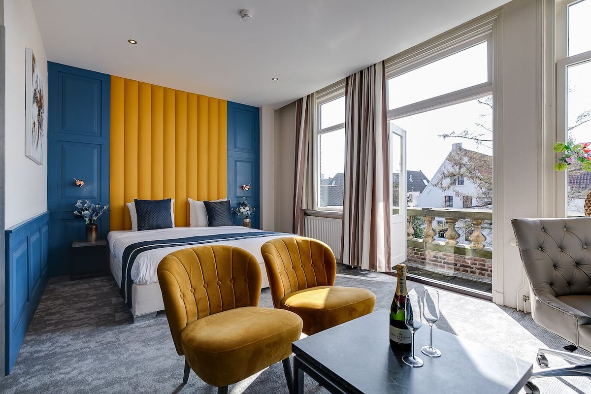 A hotel room with a bed, two yellow armchairs, a table with a bottle and glasses, and a view of buildings through large windows.