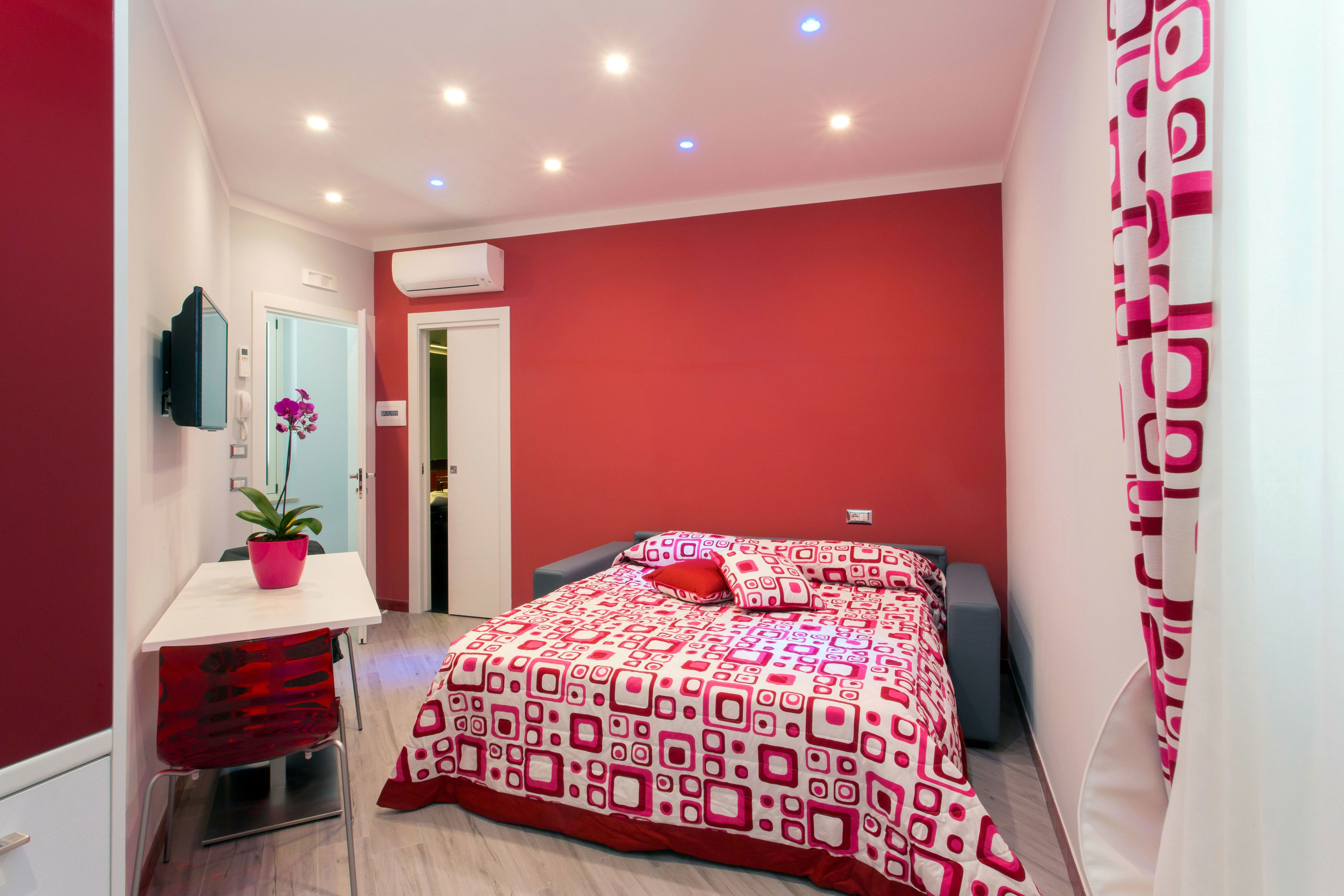 A modern bedroom with a red and white patterned bedspread, a small bedside table with a plant, and a mirror on the wall.