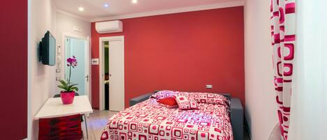 A modern bedroom with a red and white patterned bedspread, a small bedside table with a plant, and a mirror on the wall.