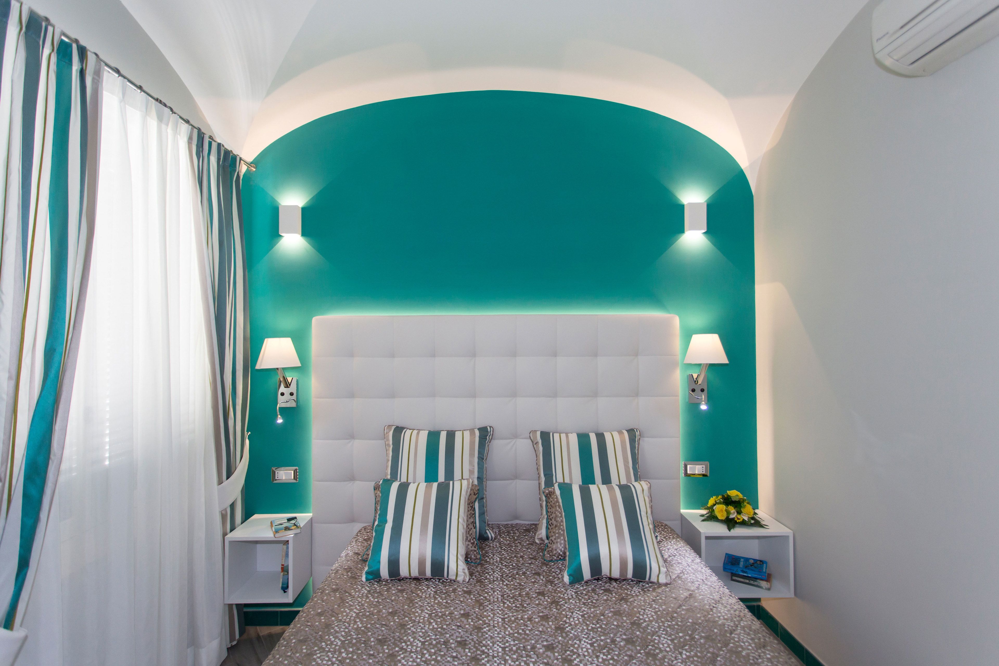 A bedroom with a teal accent wall, a tufted headboard, two bedside tables with lamps, and striped pillows.