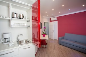 Una cocina moderna con un lavamanos integrado, armarios blancos y electrodomésticos de acero inoxidable. En la sala de estar, hay una mesa con marco rojo, una planta en maceta y un sofá gris.