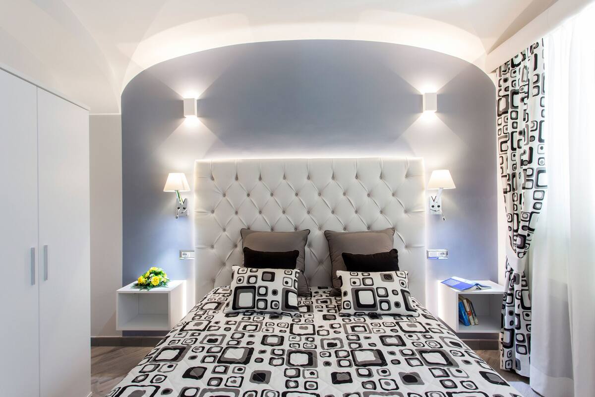 A bedroom with a tufted headboard, two bedside tables with lamps, patterned bedding, and a small bedside table with a book and a vase of flowers.