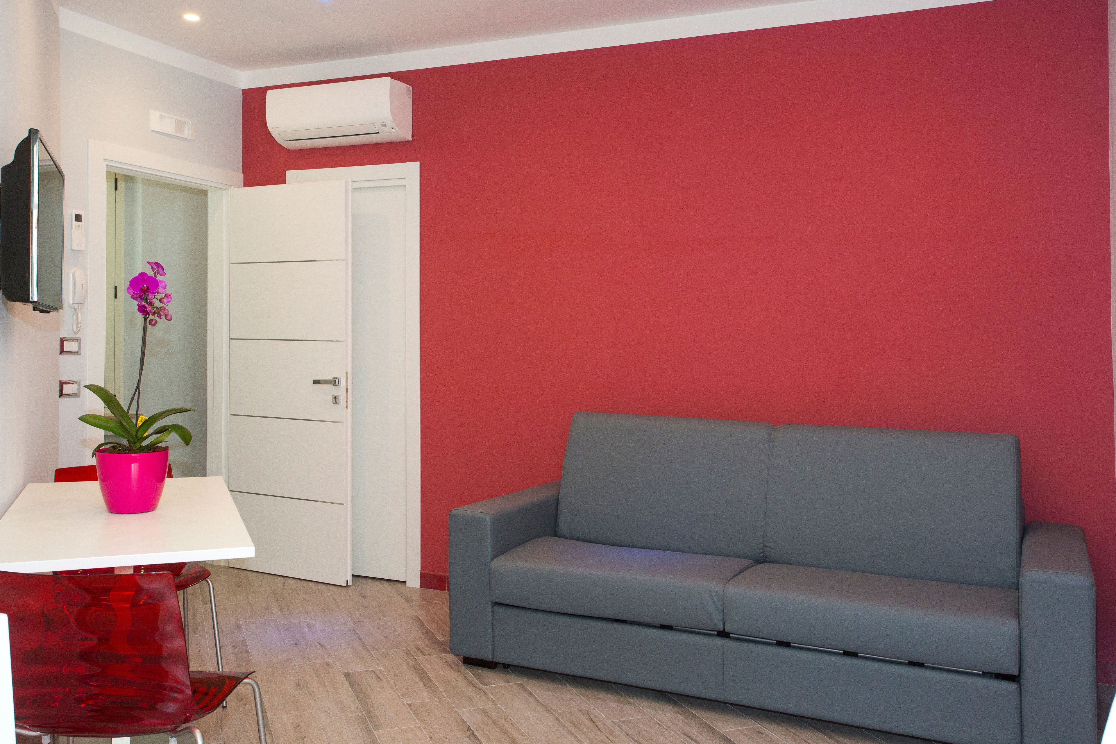 A room with a grey sofa, a white table with a pink flower pot, and a red chair.