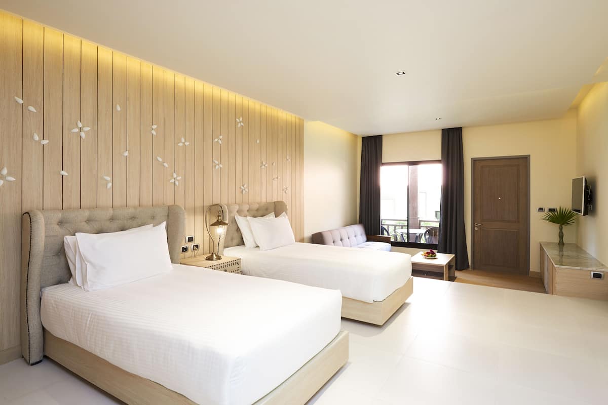 A hotel room with two beds, a wooden headboard, a bedside table with a lamp, a small table with a flower, and a TV on a stand.