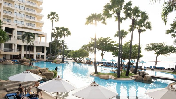 A resort pool area with lounge chairs and umbrellas, surrounded by palm trees and a large building in the background. - Garden Sea View Resort (Pattaya)