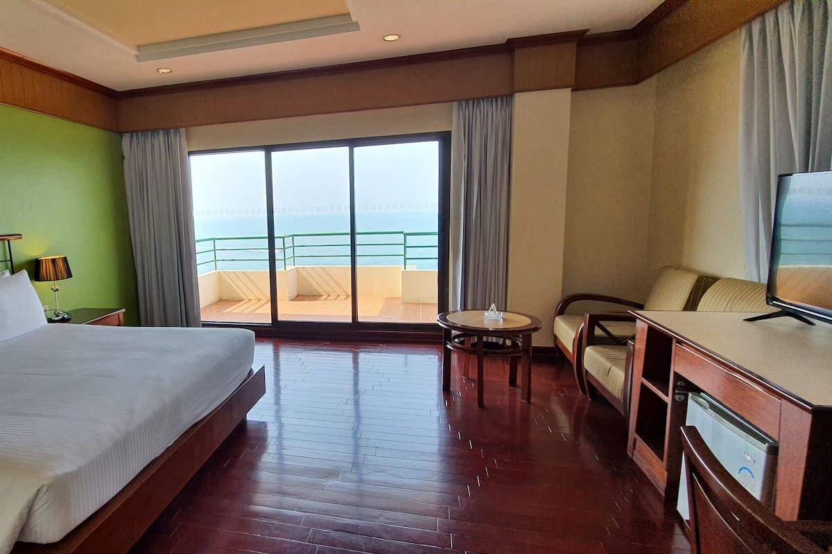 A hotel room with a large bed, a desk with a TV, a chair, and a balcony with a view of the sea.