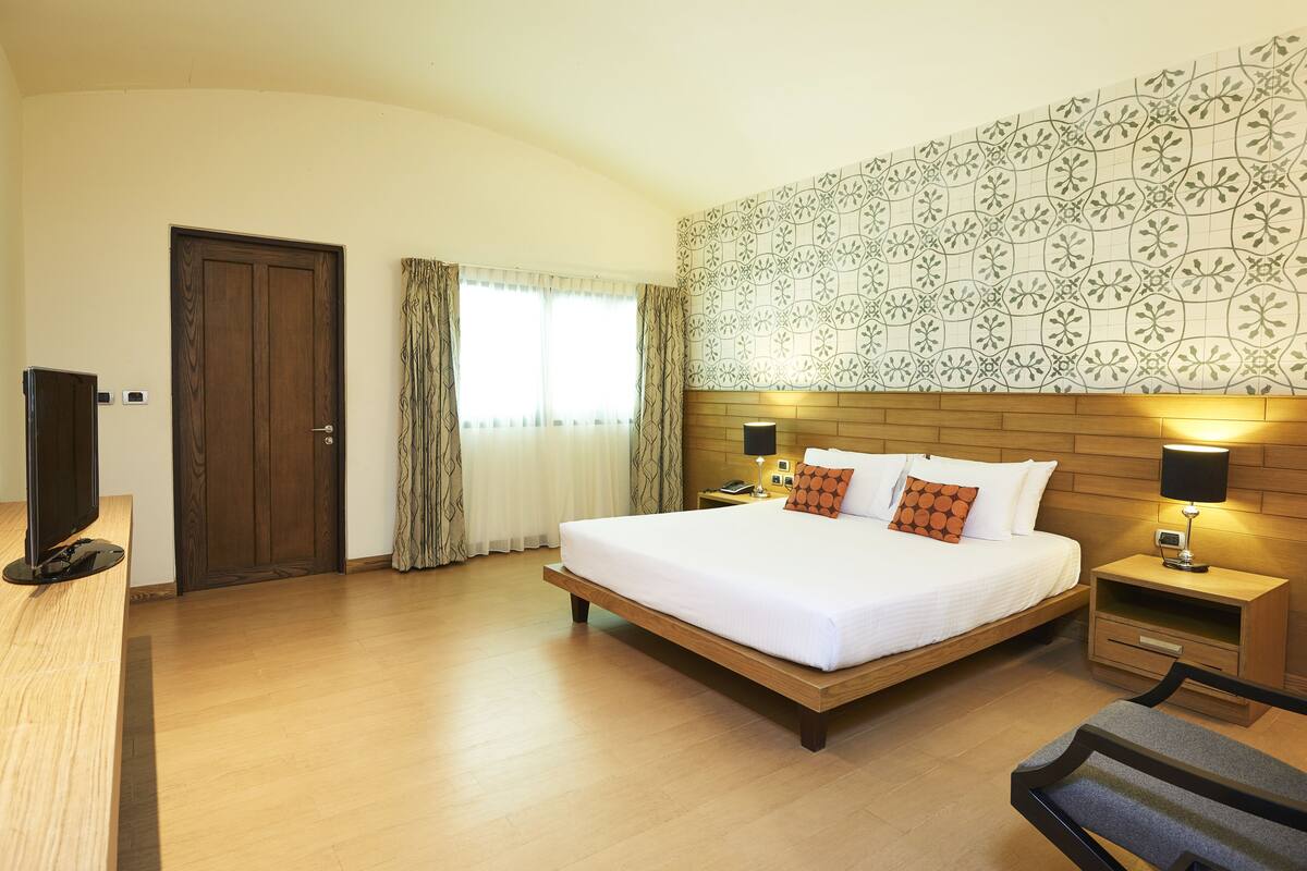 A hotel room with a large bed, wooden headboard, two bedside tables with lamps, a flat-screen TV, and a window with curtains.