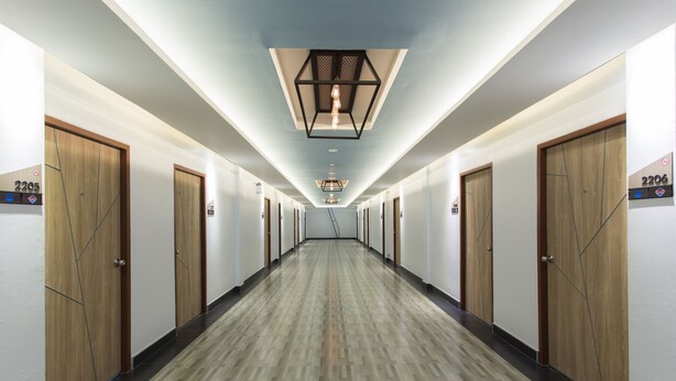 Un couloir d’hôtel avec des portes en bois, des panneaux indiquant le numéro des chambres et un luminaire suspendu au centre.