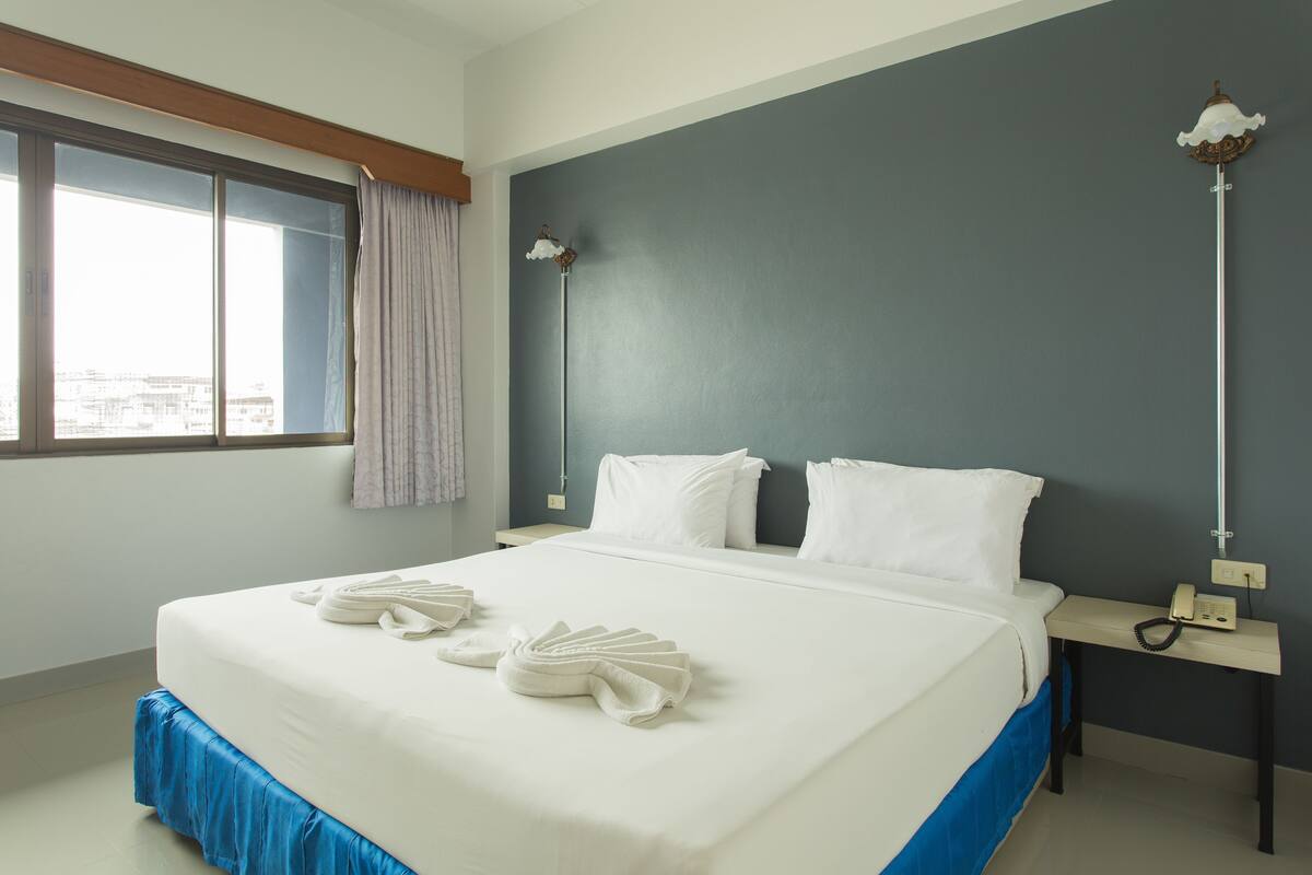 A hotel room with a large bed, a bedside table with a lamp, and a view through a window.