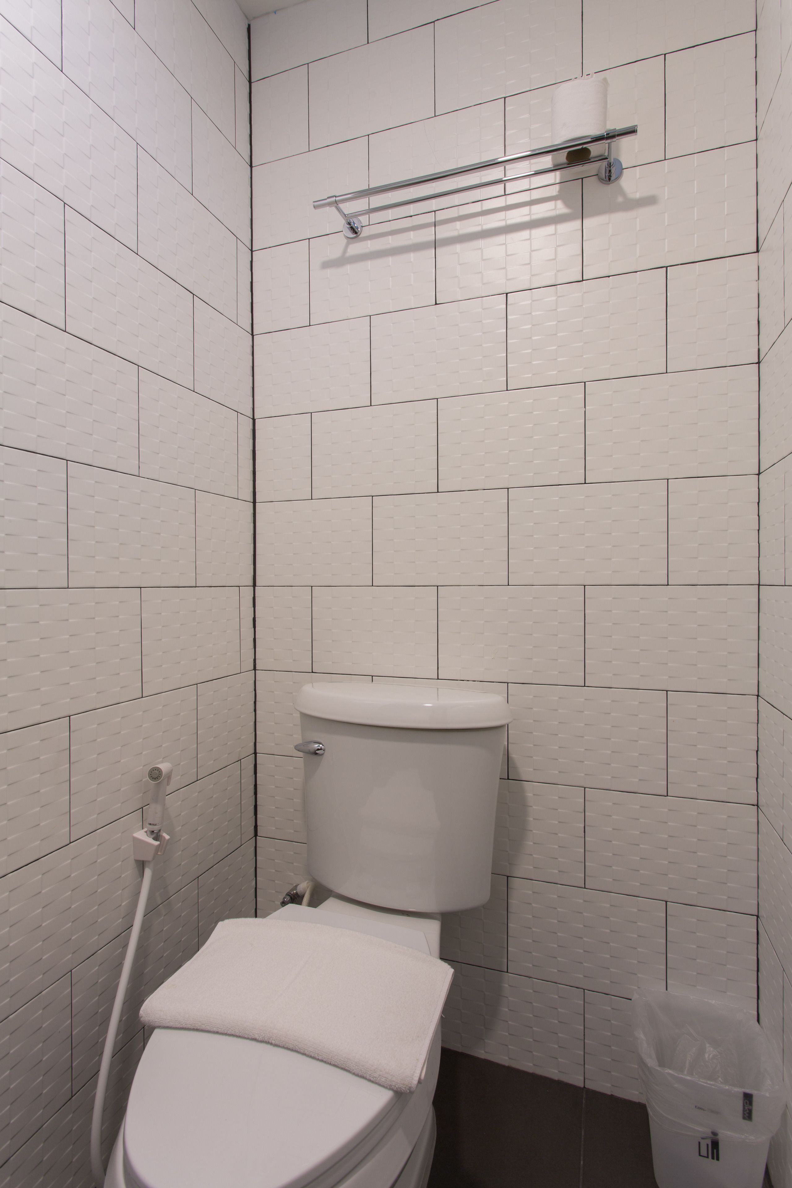 A small, white-tiled bathroom with a toilet, a towel rack, and a trash bin.