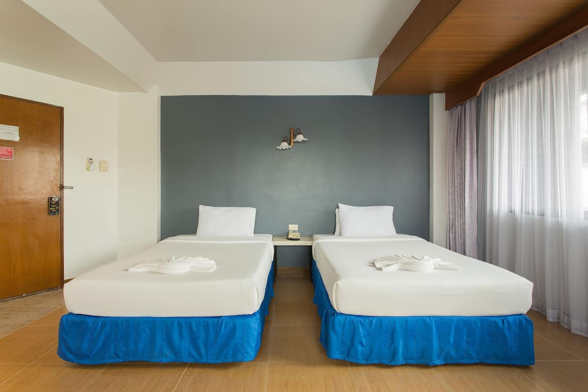 A hotel room with two beds, a wooden ceiling, a blue accent wall, a nightstand, and a window with curtains.
