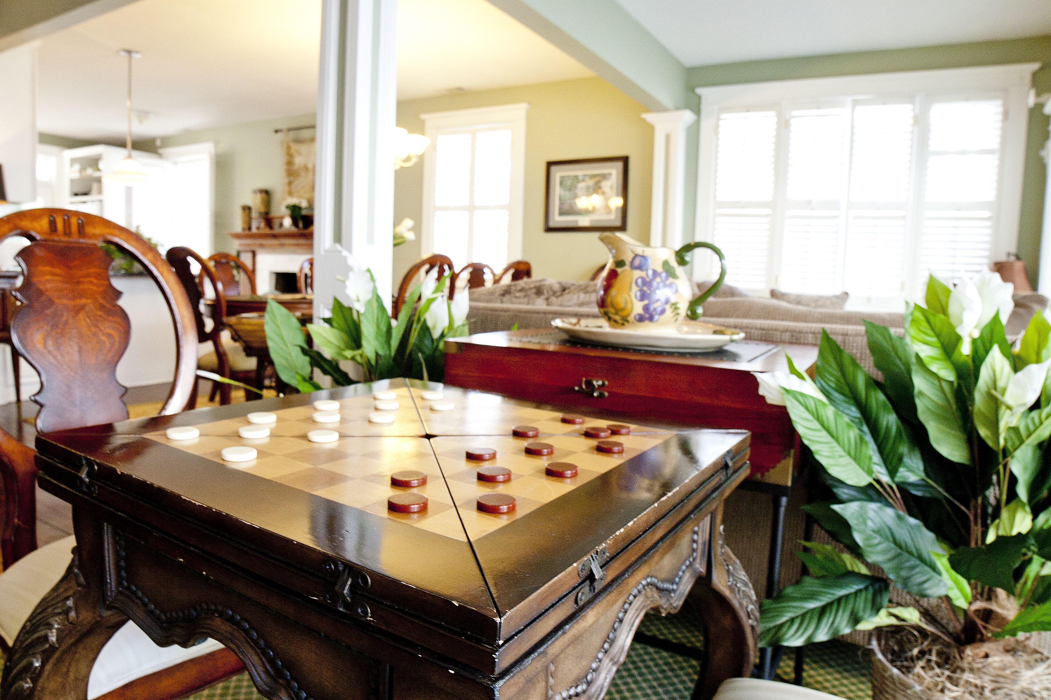 A checkered game board on a wooden table in a room with a dining area, a sofa, and potted plants.