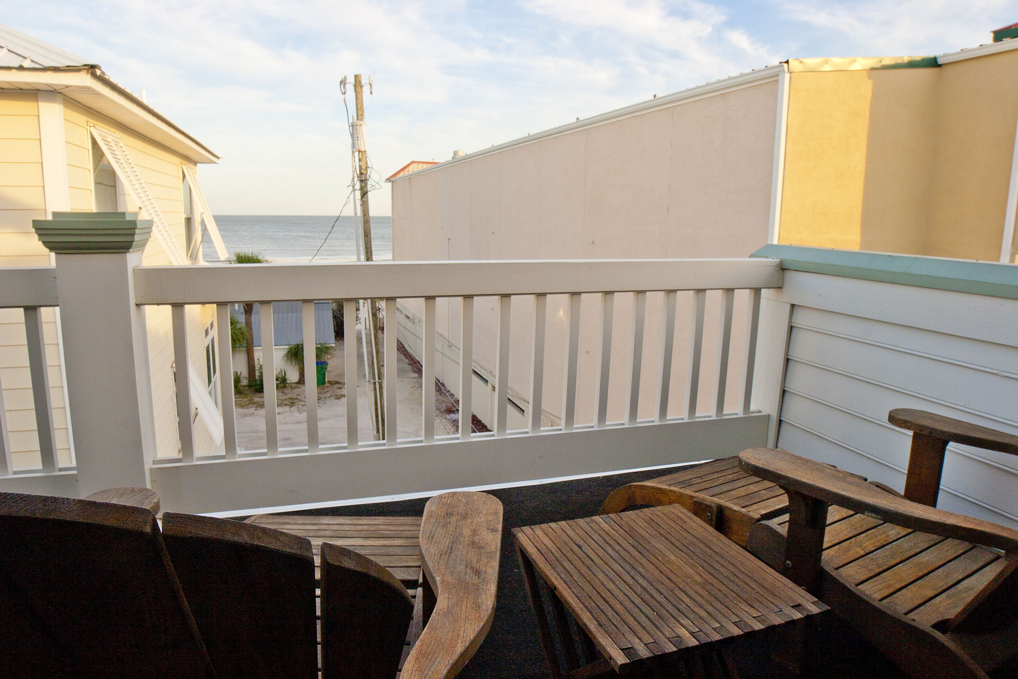 Een houten terras met een tafel en stoelen, met uitzicht op een strand en een wit gebouw.