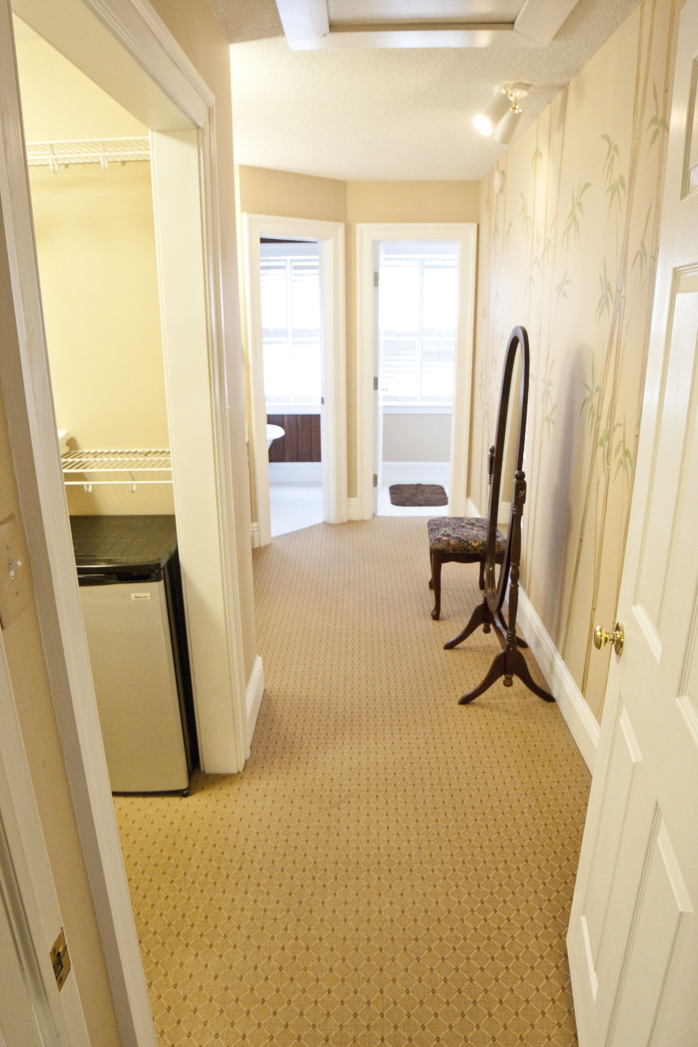 A hallway with a patterned carpet, a chair, a mirror, and a door leading to another room.