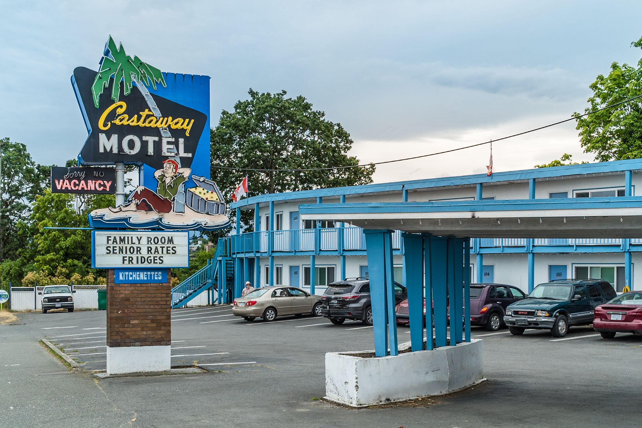 A motel with a blue and white exterior, a sign indicating family rooms, senior rates, and fridges, and a 'vacancy' sign.