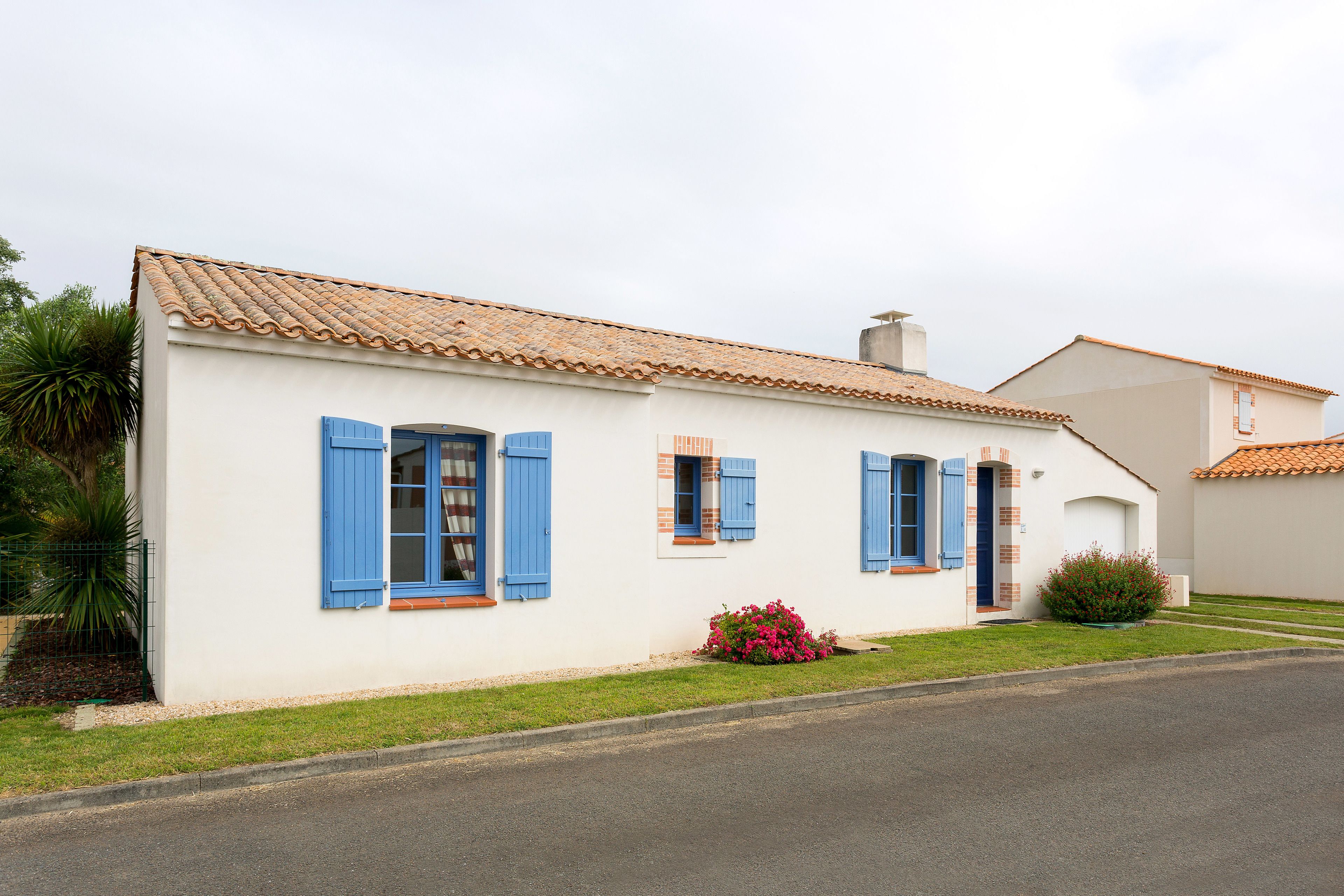 A two-story building with blue shutters, a tiled roof, and a small garden with pink flowers.