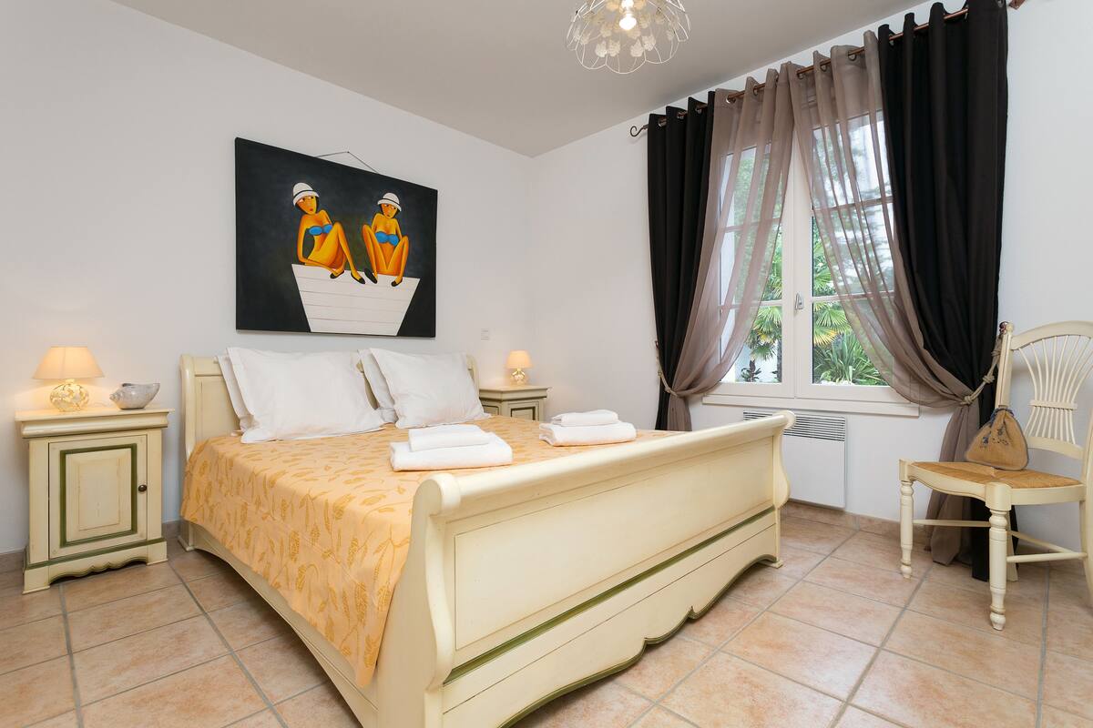 A bedroom with a bed, two bedside tables, a chair, a window with curtains, and a painting on the wall.