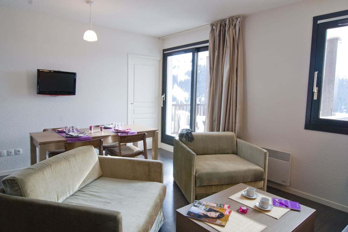 A hotel room with a sofa, armchair, dining table, and a TV mounted on the wall.