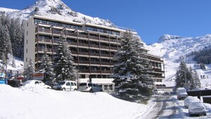 Ein mehrstöckiges Hotel mit Balkonen, umgeben von schneebedeckten Bergen und Bäumen.