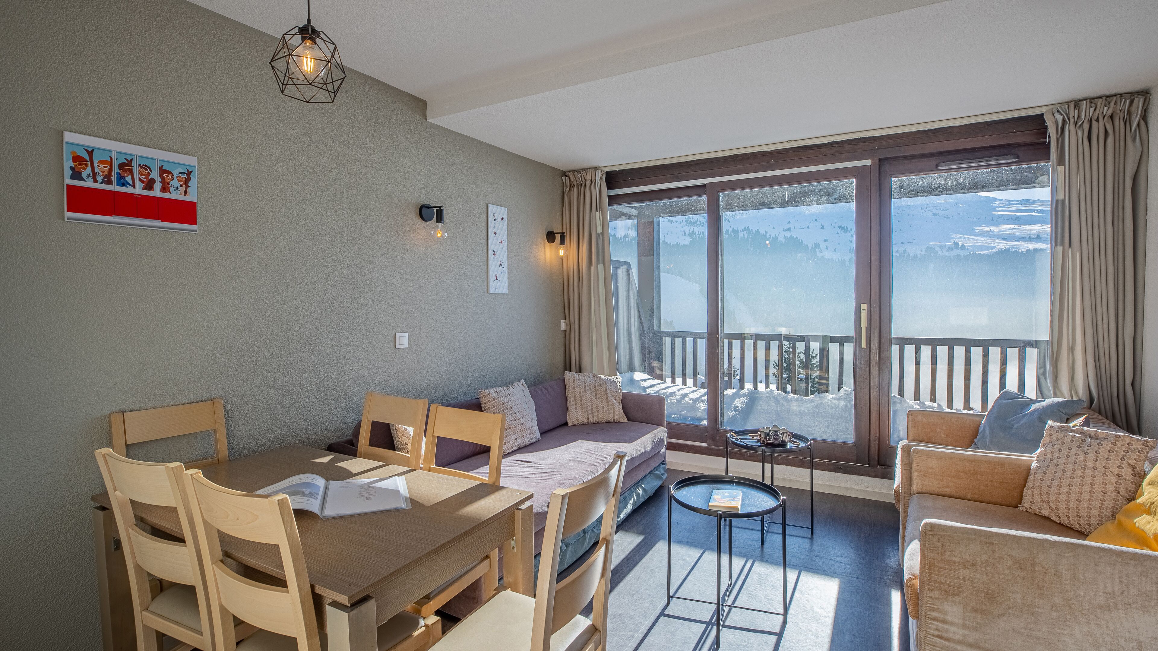 Ein gemütliches Wohnzimmer mit großem Fenster, einem Essbereich mit Holztisch und einer Wandleuchte.