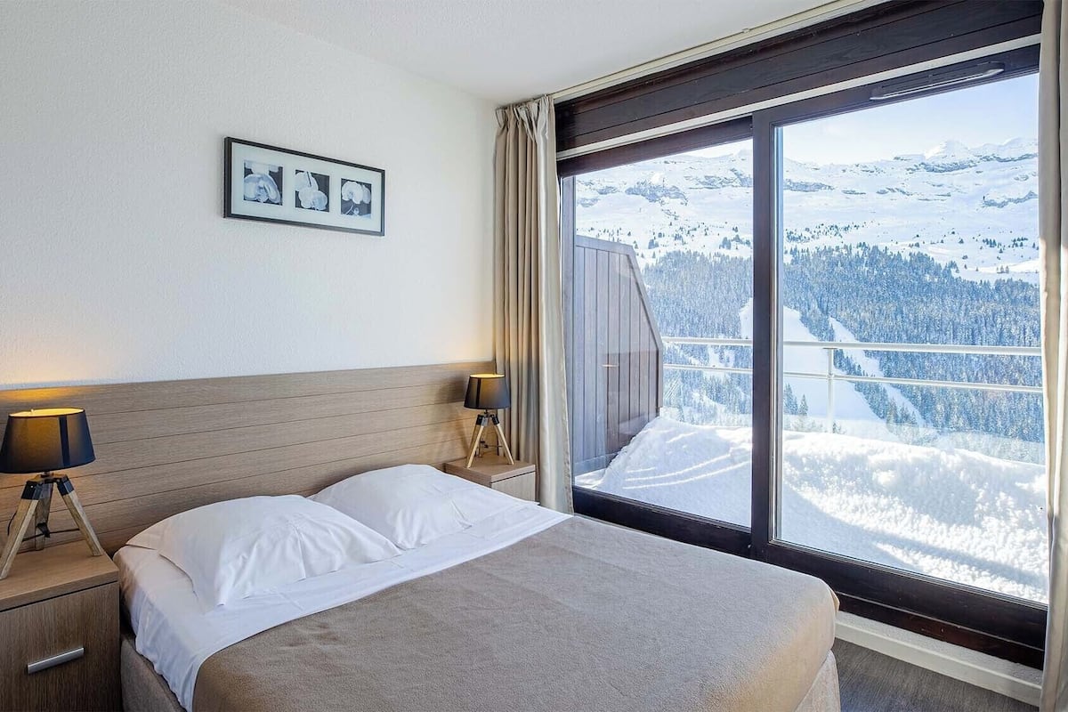 A hotel room with a bed, two bedside lamps, a framed picture on the wall, and a large window with a snowy mountain view.