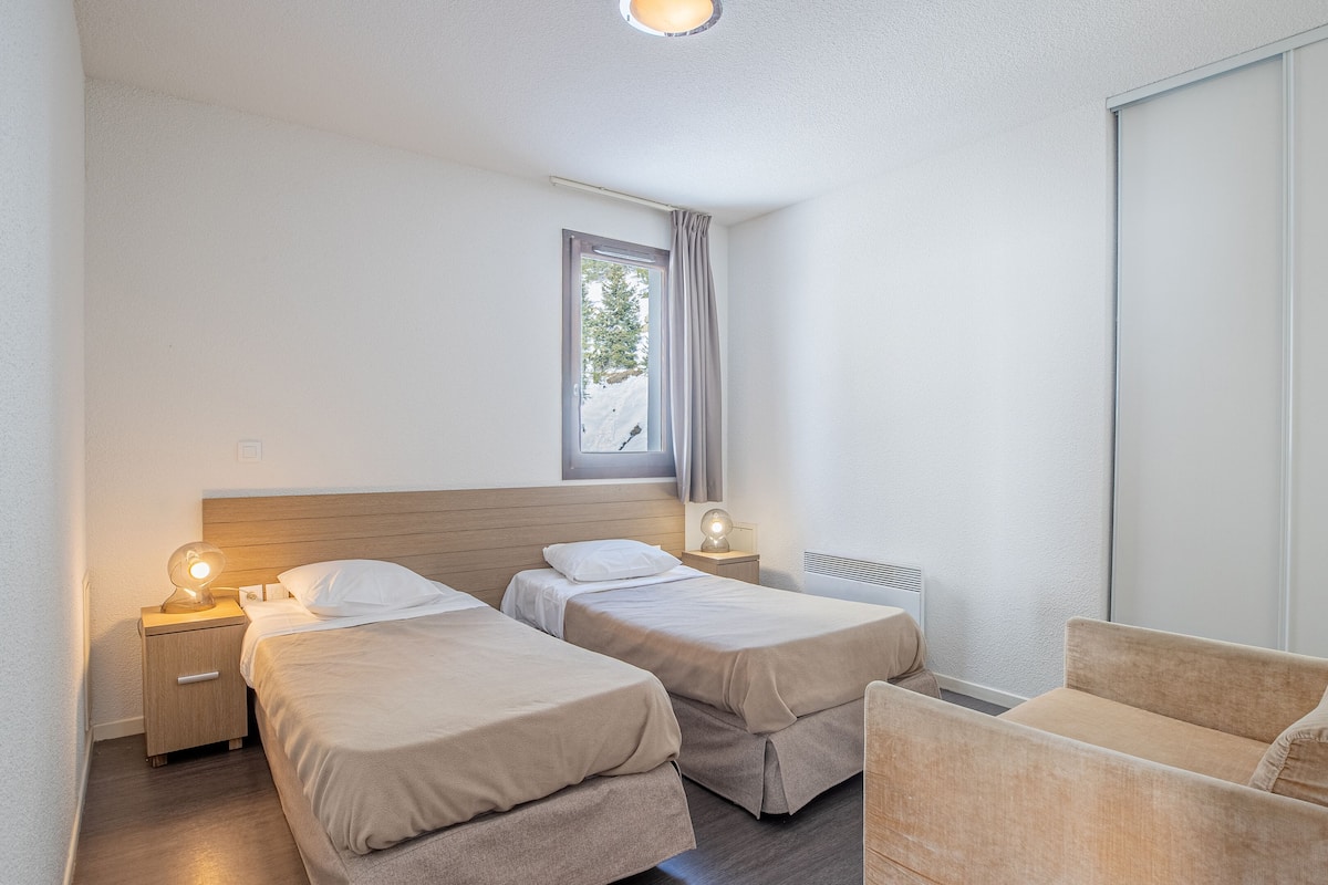 A hotel room with two single beds, a wooden headboard, bedside lamps, a window with curtains, a radiator, and a sofa.