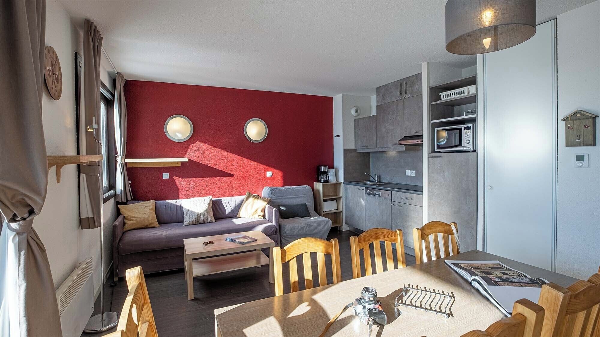 A compact living space with a red accent wall, a kitchenette, a dining area with a table and chairs, and a sofa.