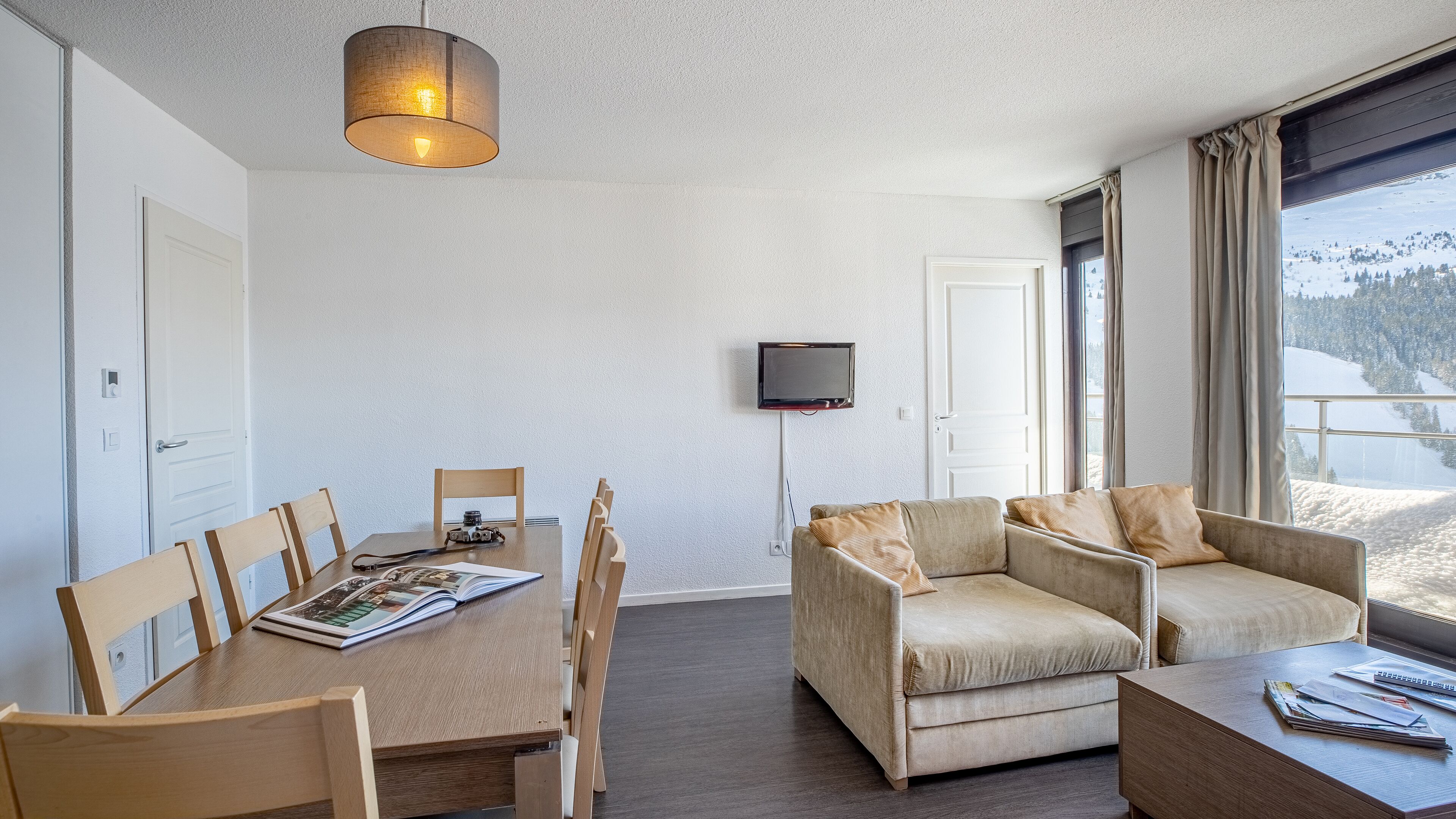 Un salon moderne comprenant une table à manger, deux canapés, un téléviseur fixé au mur et une fenêtre donnant sur un paysage enneigé.