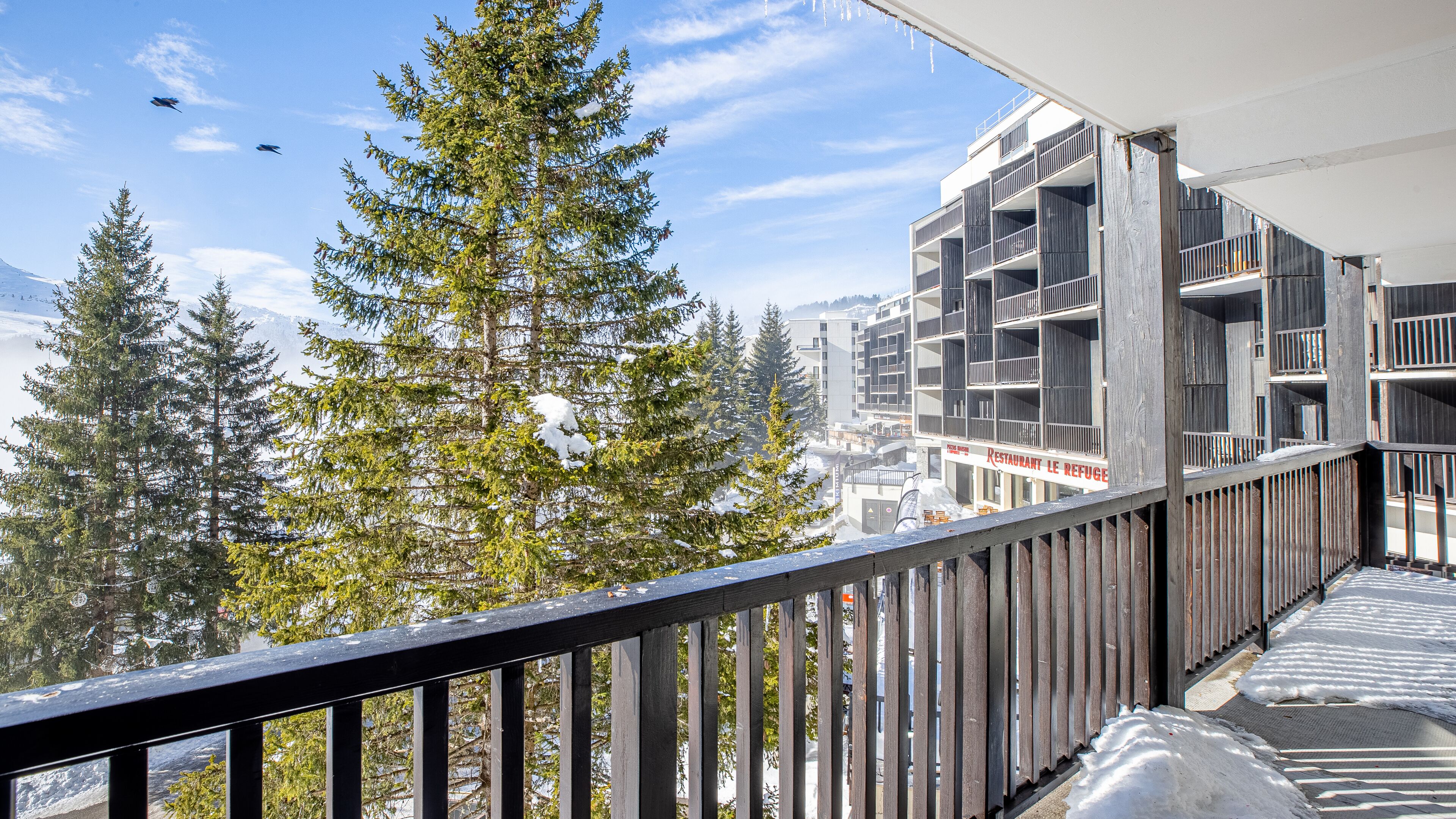 A balcony with a view of snow-covered trees and a building with a restaurant named "Restaurant Le Refuge".