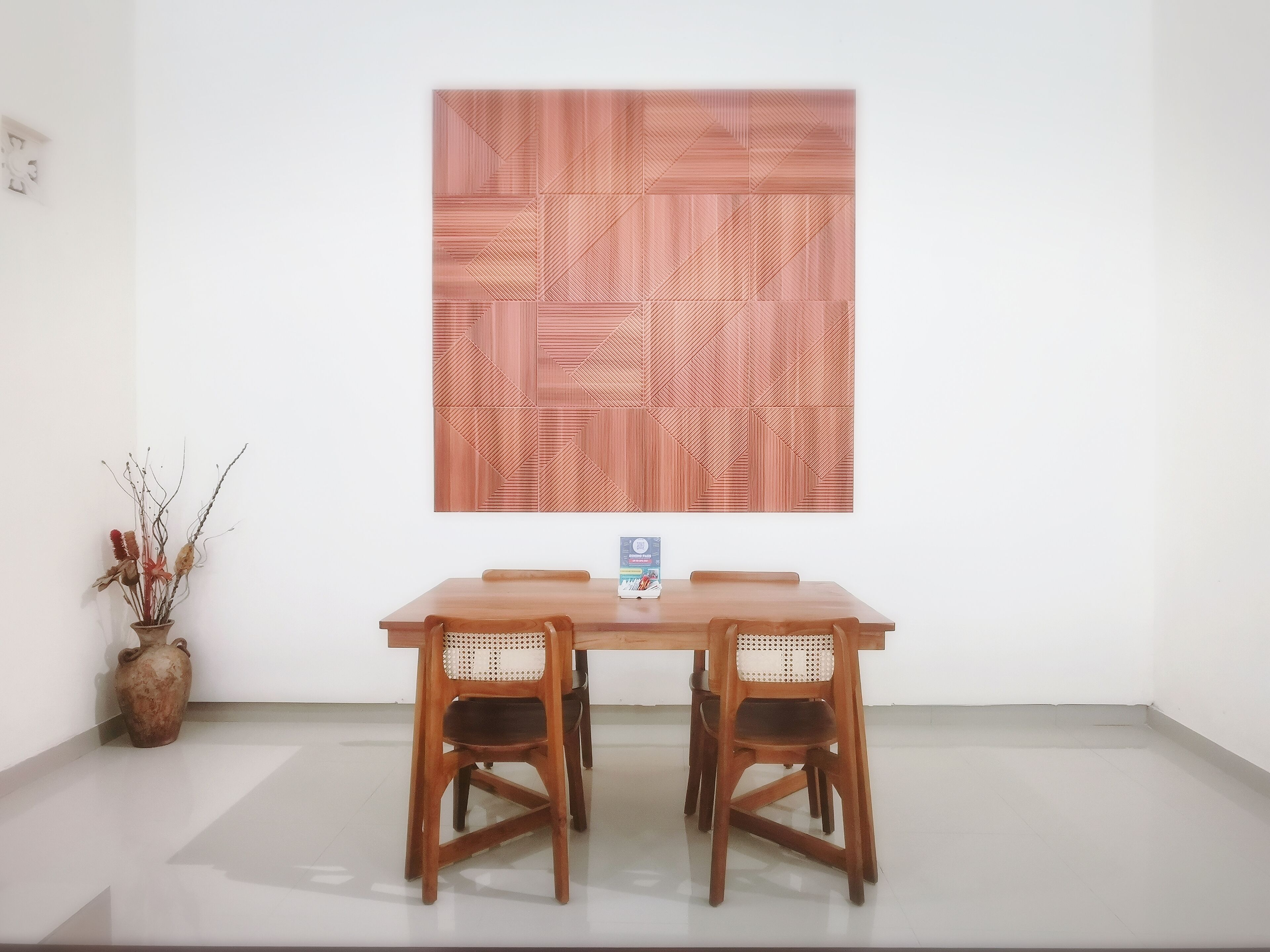 A dining area with a wooden table and chairs, a vase with dried plants, and a large geometric wall art piece.