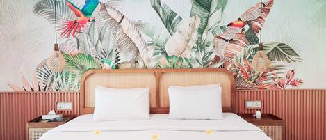 A bedroom with a large bed, two white pillows, a colorful bedspread, and a mural of tropical leaves and birds on the wall.