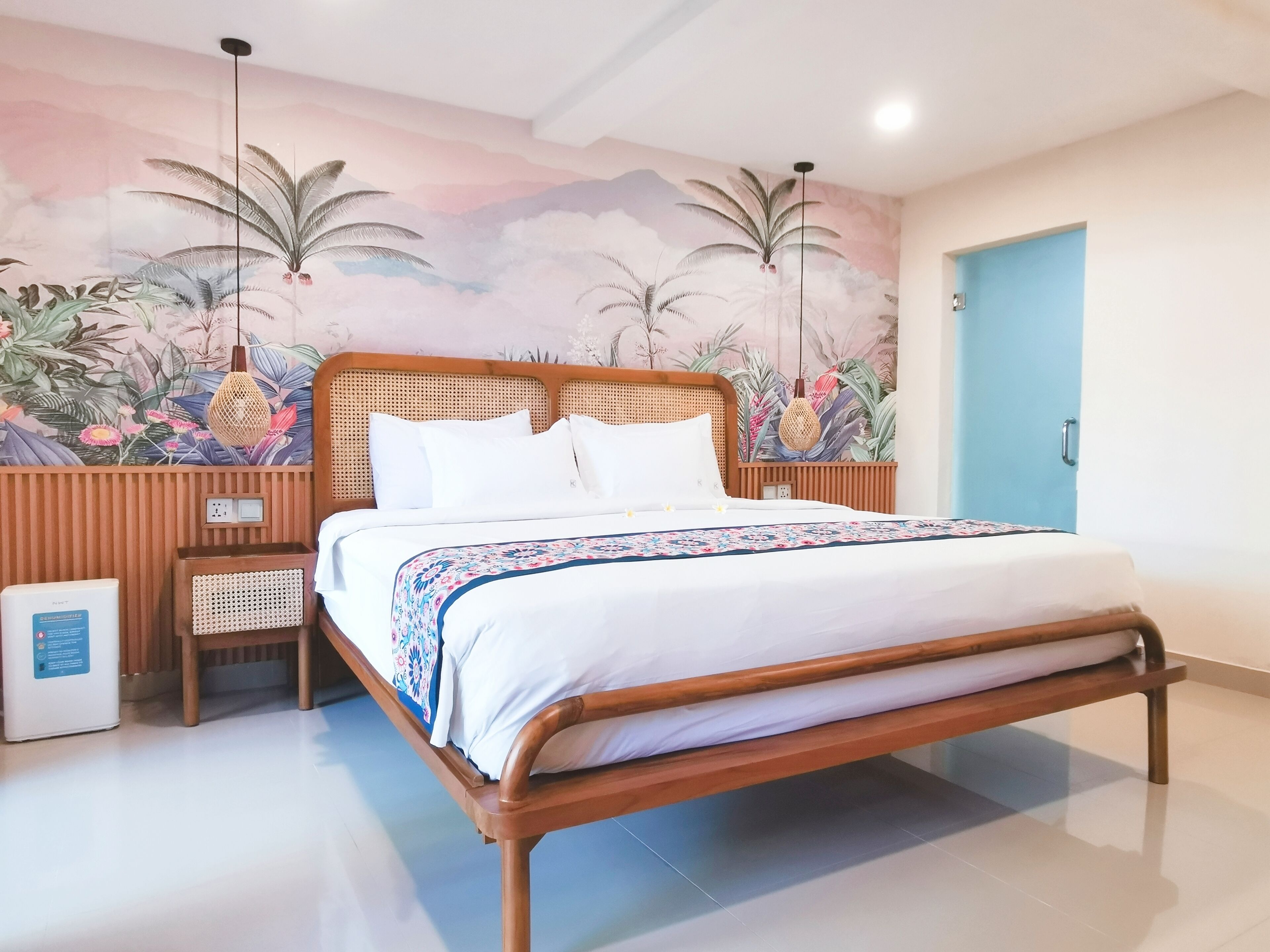 A neatly made bed with white linens and a floral-patterned blanket, a wooden headboard, and a wall mural of tropical foliage.