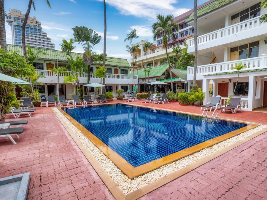 A hotel with a swimming pool surrounded by lounge chairs and umbrellas, with palm trees and a multi-story building in the background.