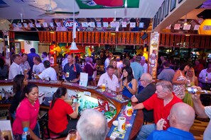 Un bar concurrido donde la gente socializa, una barra con distintos tipos de botellas y un ambiente festivo.
