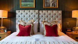 A double bed with a tufted headboard, two red pillows, and a nightstand with a lamp and radio. - Crown Inn (Chippenham)