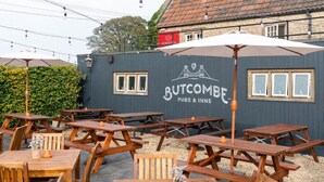 An outdoor seating area with wooden tables and chairs, a large umbrella, and a building with signage for 'Butcombe Pubs & Inns'. - Crown Inn (Chippenham)