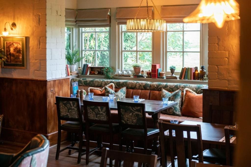 A dining area with a wooden table, chairs, and a bench with cushions. There are windows with plants and books on the windowsill.