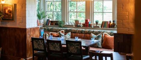 A dining area with a wooden table, chairs, and a bench with cushions. There are windows with plants and books on the windowsill.