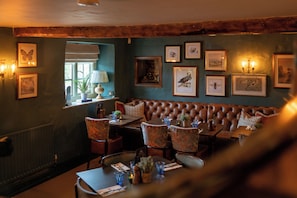 A cozy restaurant interior with a leather booth seating area, framed artwork on the walls, and a window with a view. - Crown Inn (Chippenham)