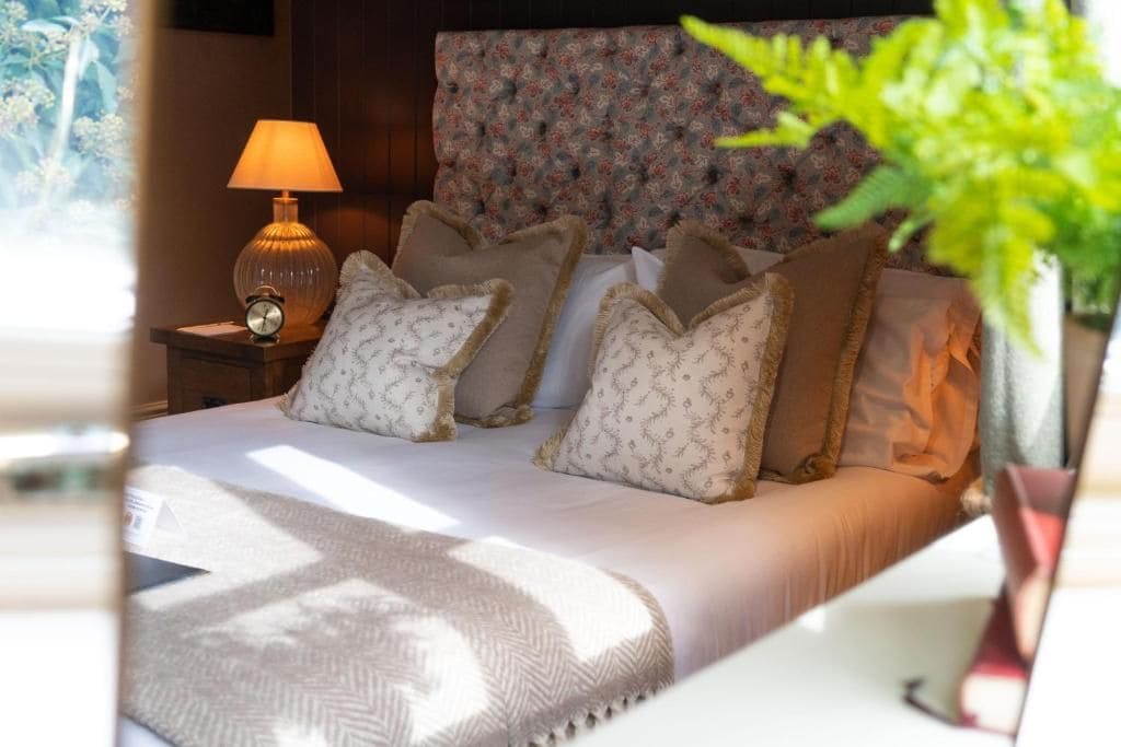 Una cama bien hecha con almohadones estampados, una lámpara de noche y un reloj en la mesita.