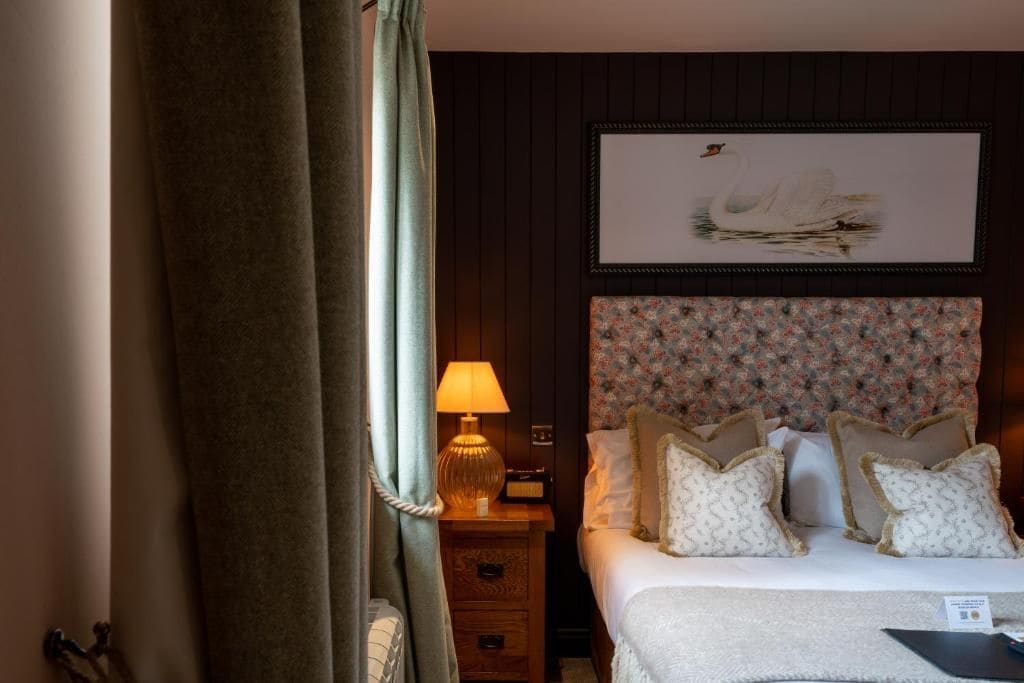A hotel room with a bed, a nightstand with a lamp, a framed picture on the wall, and a curtain partially drawn.