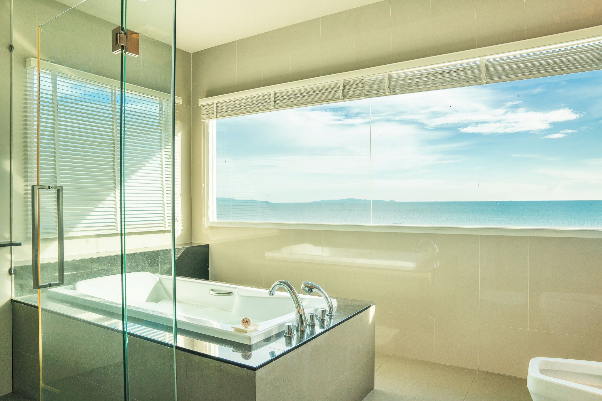 A modern bathroom with a large bathtub, a glass-enclosed shower, and a view of the sea through a large window.
