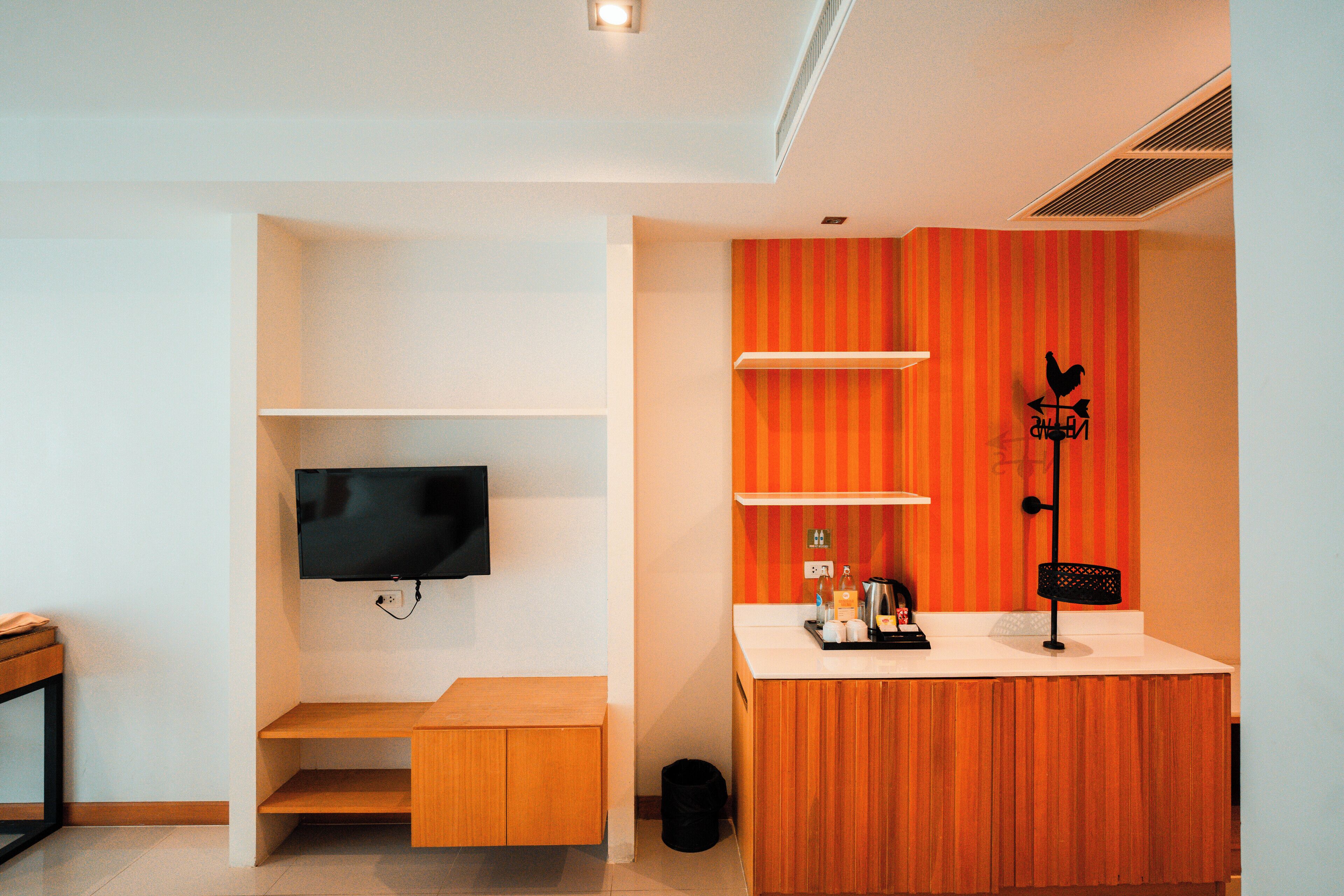 A compact living space with a flat-screen TV mounted on the wall, a wooden bench, a small table with a kettle, and a decorative wall piece.