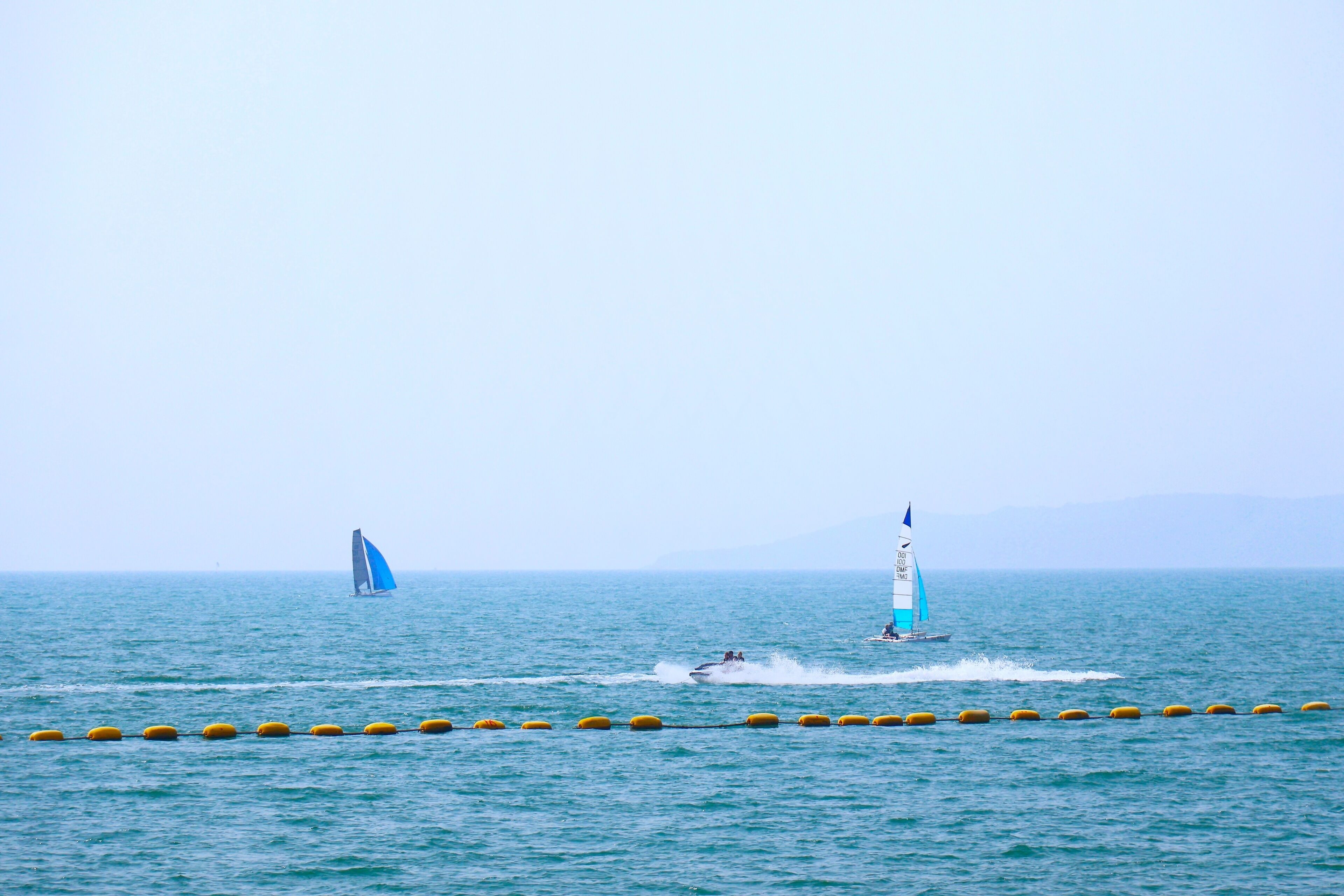 Une mer calme, avec des bouées et deux voiliers.