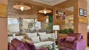 A modern hotel lobby with a beige sofa, a purple armchair, and a glass coffee table. There are decorative wall panels and a large mirror. - Boudl Al Maidan (Hafr Al Batin)