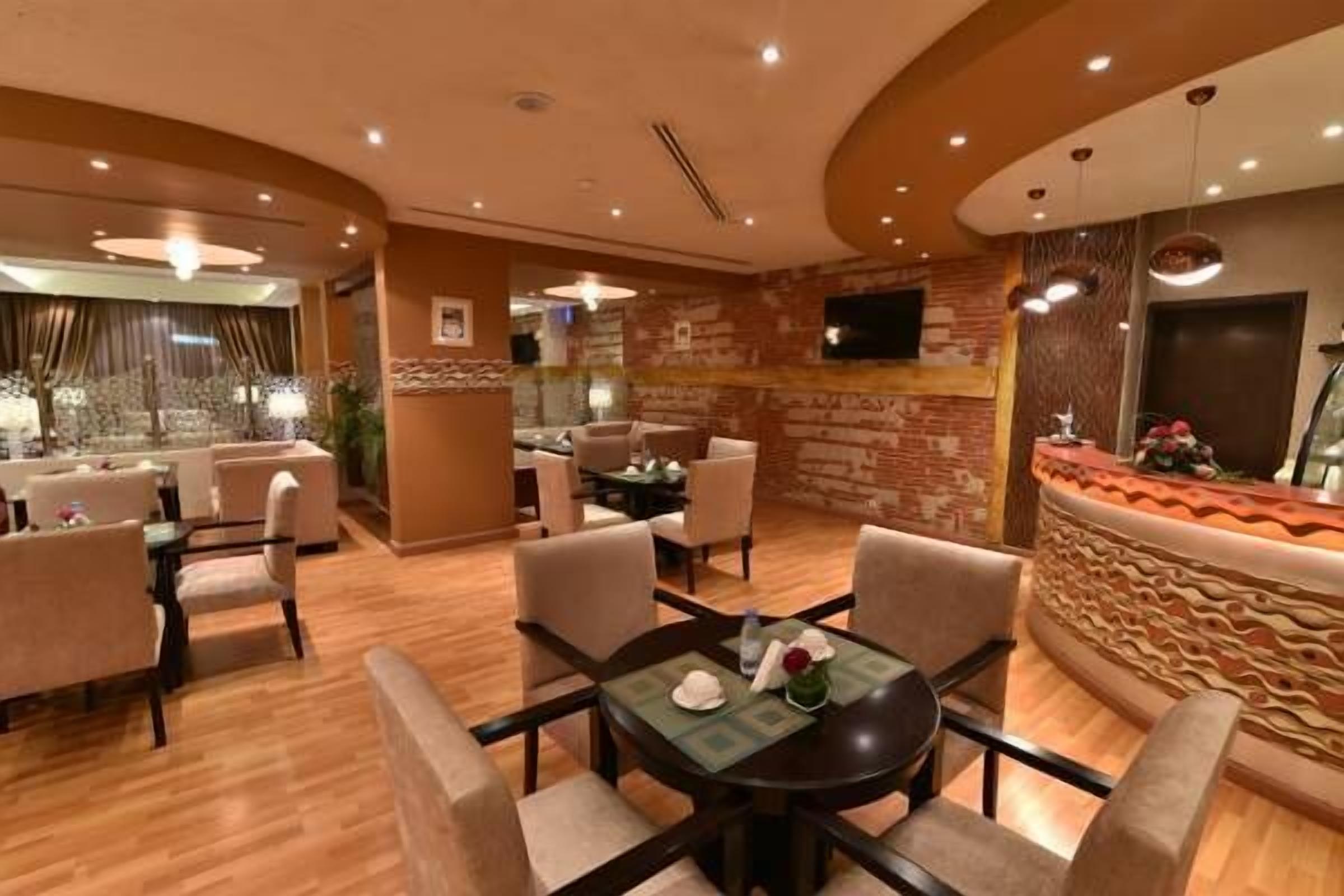 Un vestíbulo de hotel con piso de madera, mesas redondas y sillas color beige. Hay una recepción con un muro decorativo y un televisor montado encima.