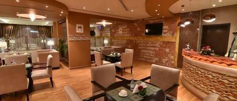 Un vestíbulo de hotel con piso de madera, mesas redondas y sillas color beige. Hay una recepción con un muro decorativo y un televisor montado encima.