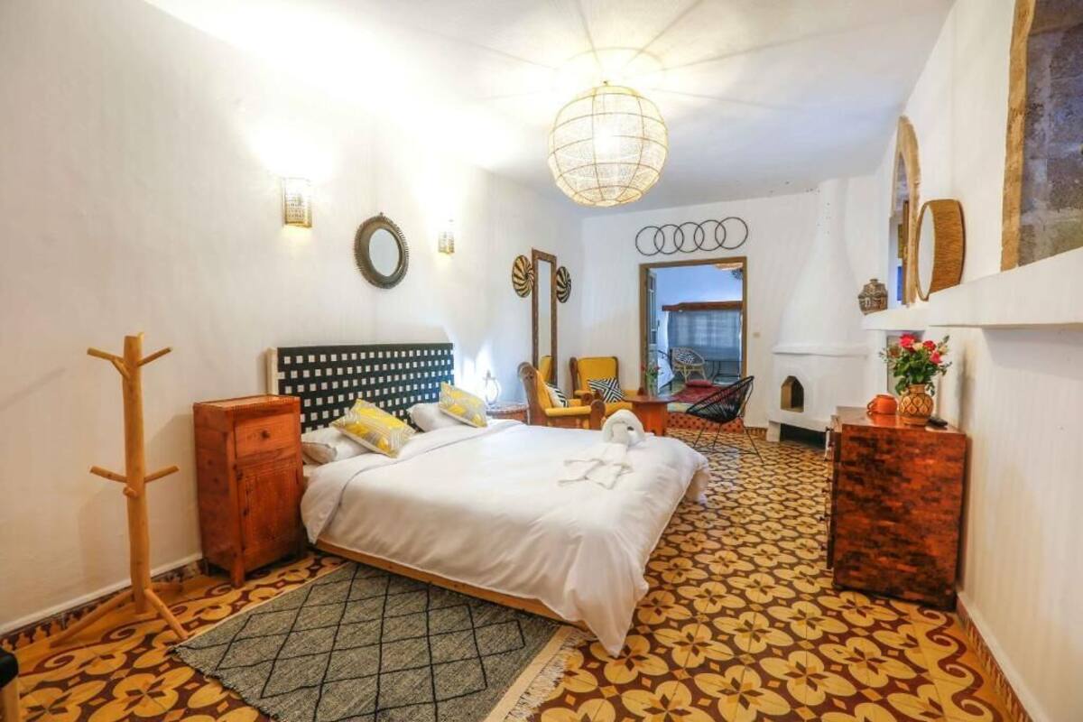 A hotel room with a bed, a wooden side table, a chair, a mirror, and a patterned floor.